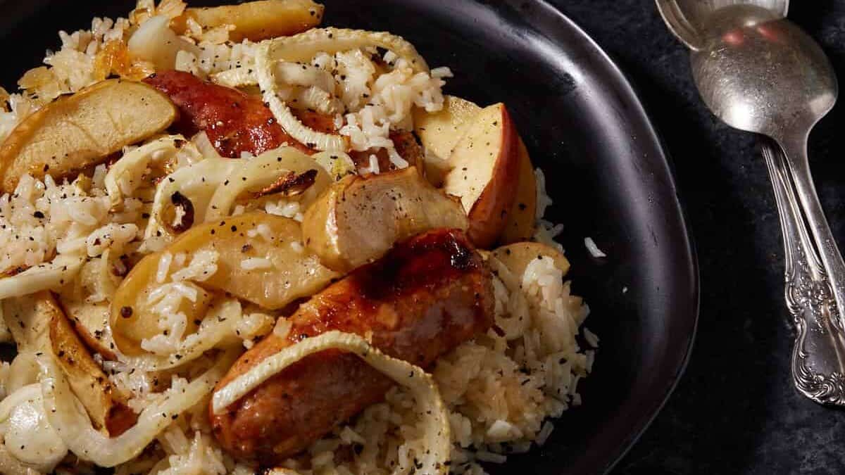 A black platter filled with sausages and apples next to two serving spoons.