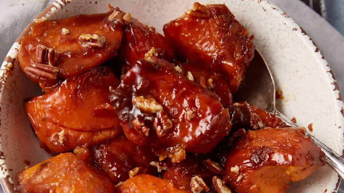 A bowl of glazed sweet potatoes garnished with chopped pecans. The sweet potatoes are caramelized and served in a rustic, white ceramic bowl placed on a gray cloth. A spoon is partially visible in the bowl.