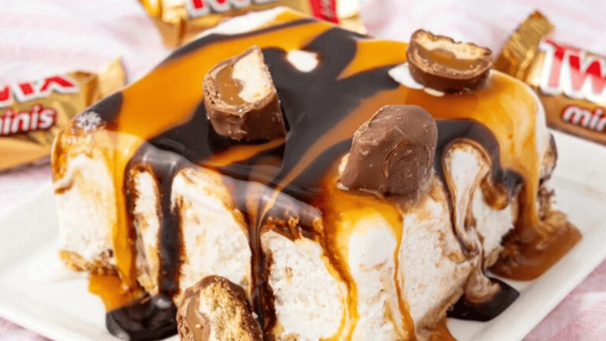 A rectangular ice cream cake topped with caramel and chocolate sauce, garnished with pieces of Twix Minis candy bars—an indulgent twist on Thanksgiving treats, with more Twix Minis in the background.