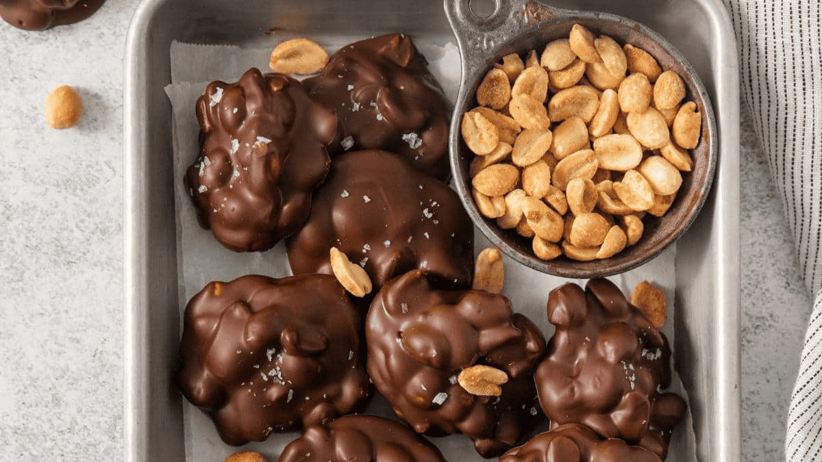 Chocolate-covered peanut clusters on a metal tray with a small bowl of salted peanuts—perfect chocolate desserts for Christmas. Some clusters are topped with peanuts and sea salt, with a striped towel partially visible at the tray’s edge.