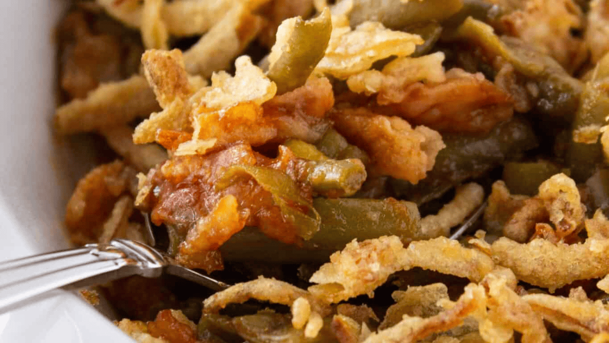 Close-up of green bean casserole, a classic among Christmas casseroles, topped with crispy fried onions—showing green beans, creamy sauce, and golden brown onion pieces, with a fork visible in the bottom left corner.