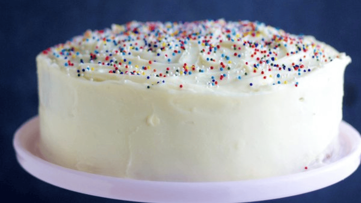 A round cake covered in smooth white frosting, reminiscent of classic desserts with cream cheese, is topped with colorful rainbow sprinkles and sits on a light pink cake stand against a dark blue background.