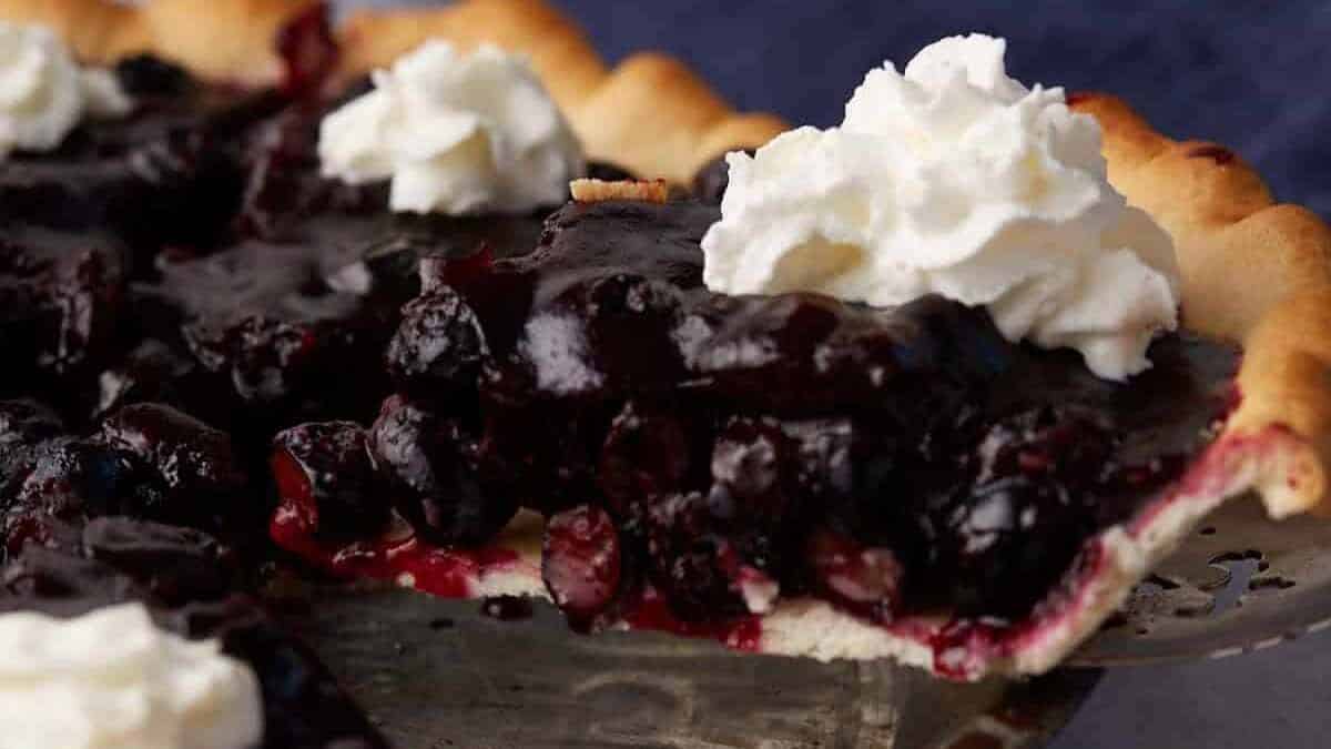 A freshly sliced blueberry pie topped with dollops of whipped cream is displayed on a metal pie dish. The deep purple filling contrasts with the golden crust, and the pie is partially cut, showcasing the juicy blueberries.