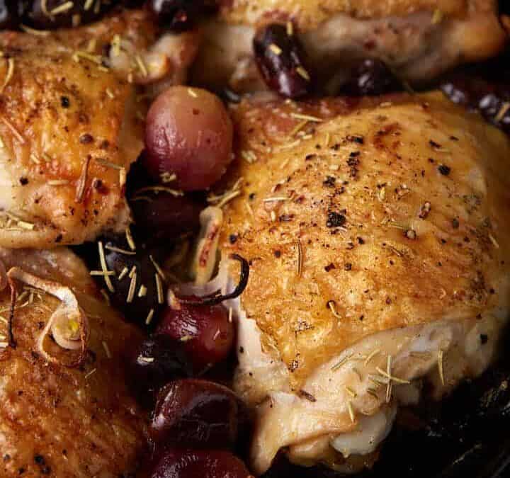 A skillet of chicken thighs with olives and grapes.