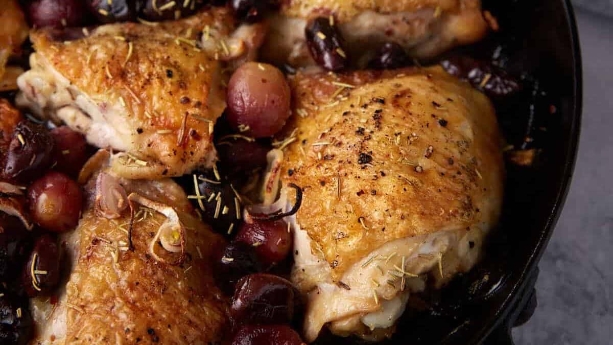 A skillet of chicken thighs with olives and grapes.