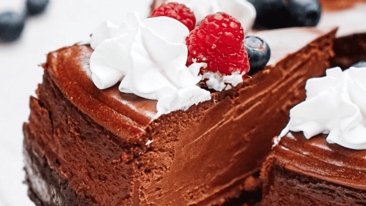 A close-up of a rich chocolate cheesecake—one of the best chocolate desserts for Christmas—topped with whipped cream, fresh raspberries, and blueberries. One slice is partially separated from the rest of the cake.