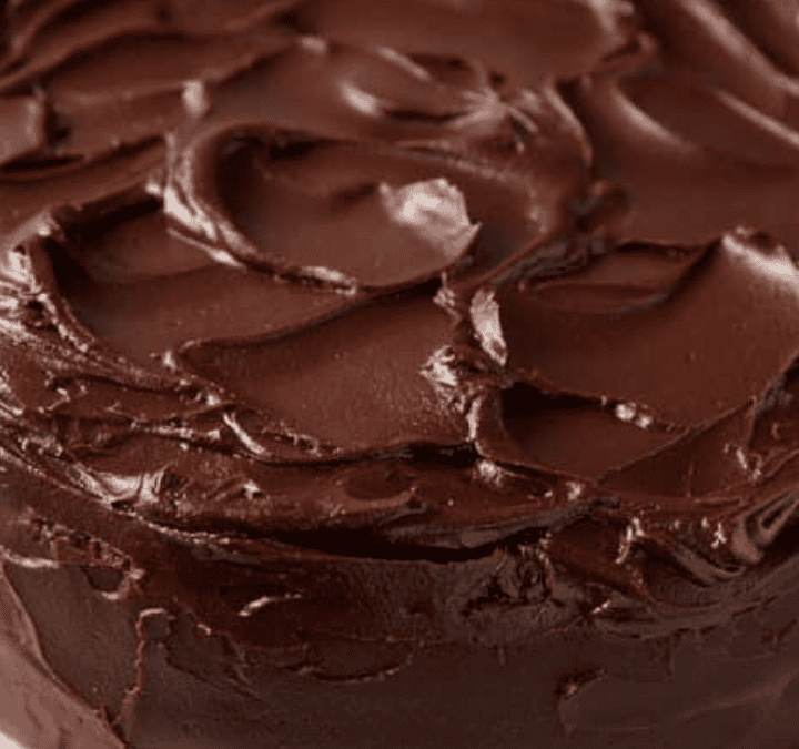 A round chocolate cake covered in thick, glossy chocolate frosting sits on a white plate. The indulgent dessert features frosting spread with textured swirls across the top and sides of the cake.