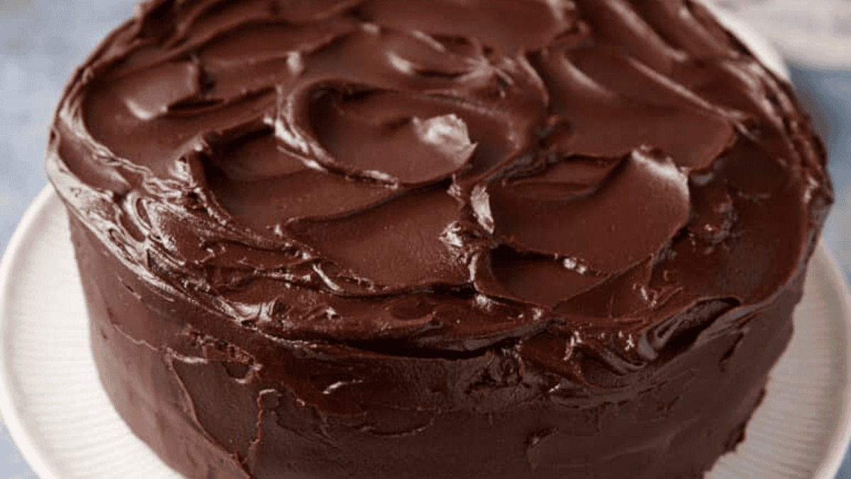 A round chocolate cake covered in thick, glossy chocolate frosting sits on a white plate. The indulgent dessert features frosting spread with textured swirls across the top and sides of the cake.