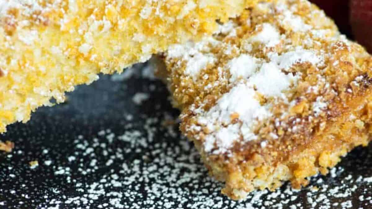 Close-up of two golden French Toast Sticks dusted with powdered sugar on a black plate. In the background, two fresh strawberries are partially visible, adding a touch of color to the presentation.