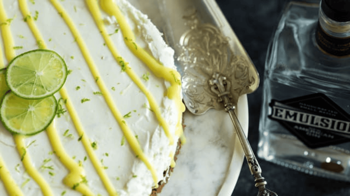 A close-up of a cheesecake dessert with white frosting, yellow drizzle, and lime slices on top, placed on a marble tray with a vintage serving utensil and a bottle labeled "EMULSION" beside it.