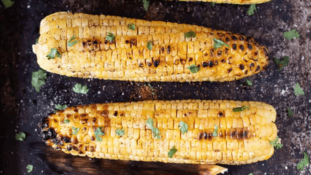 Two grilled corn on the cob, classic easy grilled foods, sprinkled with chopped cilantro and pepper. The corn boasts visible grill marks and a slightly charred appearance, resting on a dark surface.