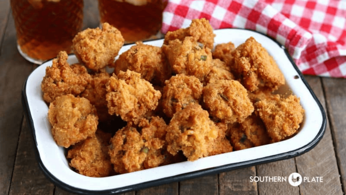 A white enamel tray filled with golden brown, quick dippable appetizers—hush puppies—sits on a wooden table, with glasses of iced tea and a red-checkered napkin in the background.