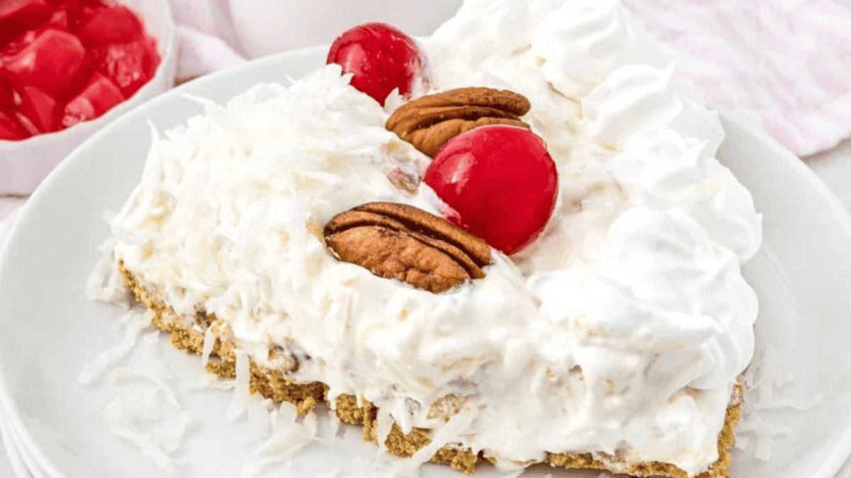 A slice of creamy coconut pie topped with whipped cream, two pecan halves, and two red cherries, served on a white plate—a festive no bake Christmas treat. A bowl of red cherries is visible in the background.