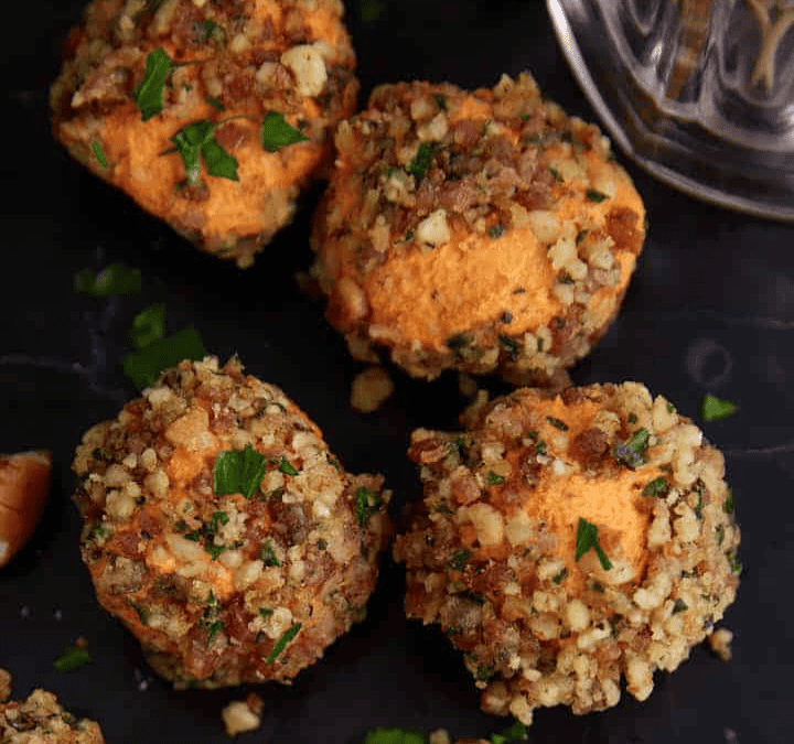 Four cheese balls coated with chopped nuts and herbs are arranged on a dark serving surface, next to a bowl of pickles and a glass mug—perfect as savory NYE party foods for an elegant appetizer presentation.