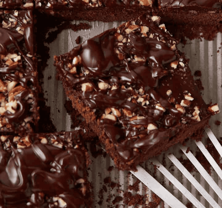 A tray of chocolate brownies, perfect for holiday cakes and cupcakes spreads, topped with chopped nuts and drizzled with chocolate, with one brownie being lifted out with a metal spatula.