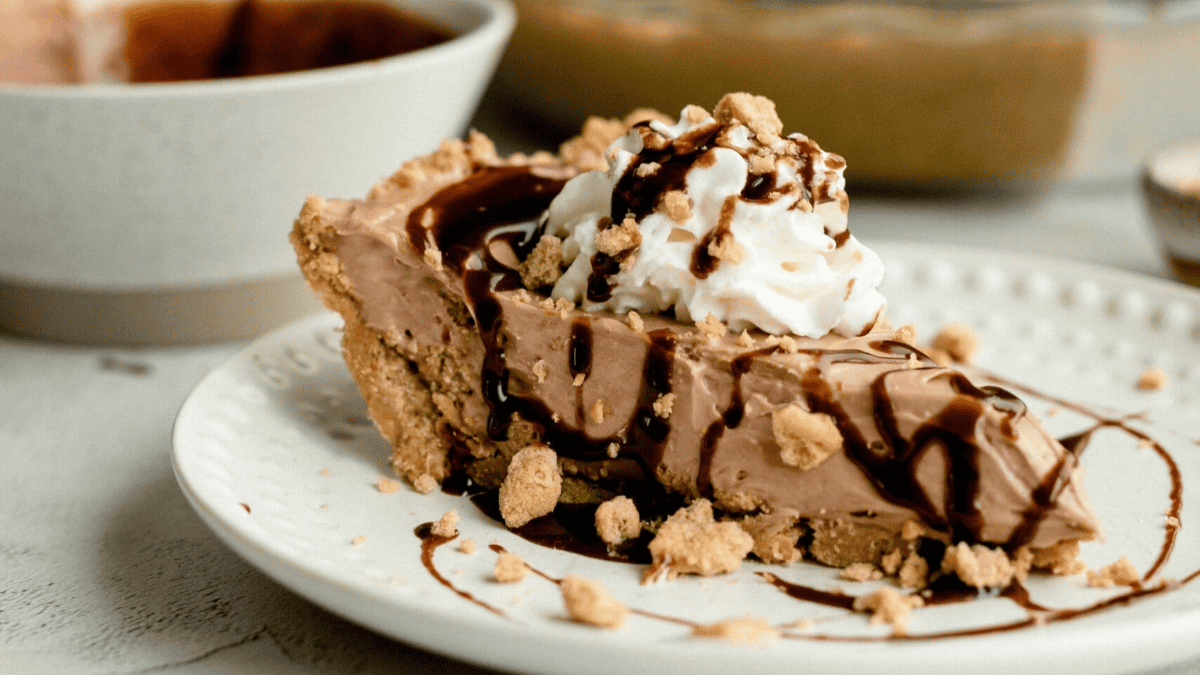 A slice of chocolate cream pie with a graham cracker crust, topped with whipped cream, chocolate syrup, and crumbled graham crackers—perfect for holiday gatherings and those searching for new christmas pie recipes—served on a white plate.