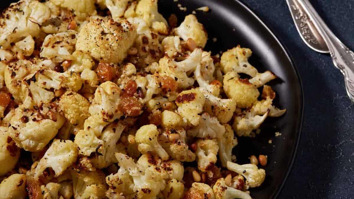 A black platter of roasted cauliflower with garlic next to serving spoons.