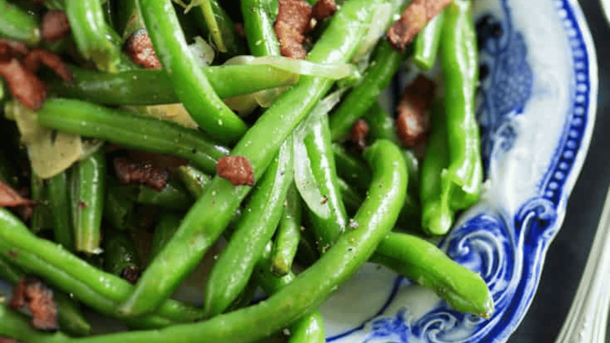 A close-up of cooked green beans mixed with pieces of bacon and sliced onions, served on a decorative blue and white plate—perfect for adding to your holiday side dishes spread.