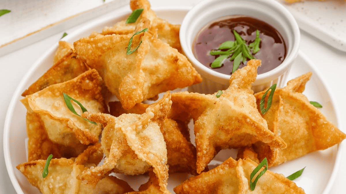 A plate of golden-brown, crispy fried wontons—perfect quick dippable appetizers—garnished with sliced green onions and served with a small bowl of dark dipping sauce on the side.