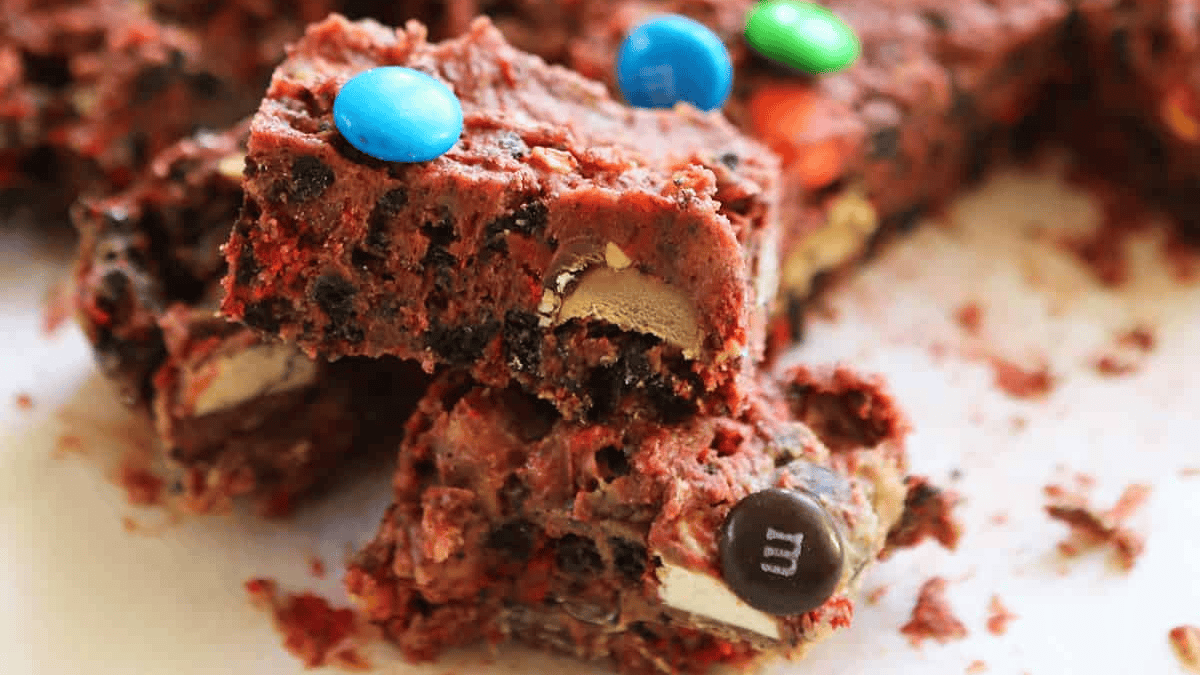Close-up of colorful candy-studded rocky road bars with chocolate, cookies, and blue and brown coated candies visible, stacked on a white surface—an irresistible no bake dessert perfect for any sweet tooth.