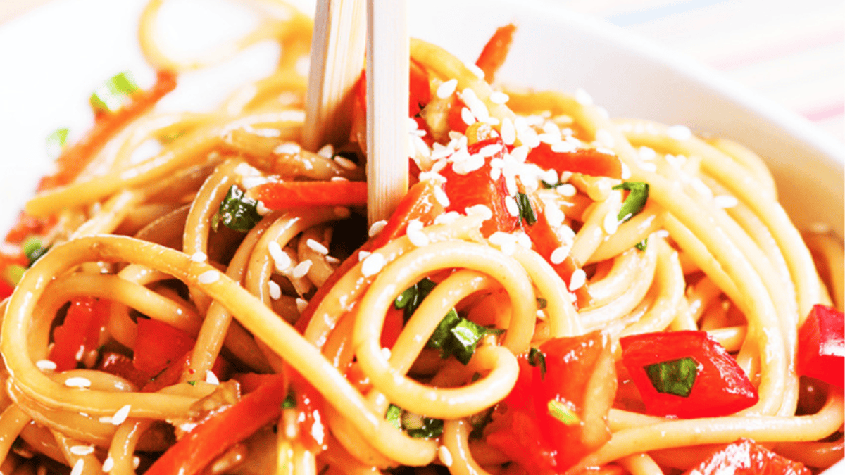 Close-up of chopsticks picking up saucy noodles mixed with red bell peppers, green herbs, and topped with white sesame seeds—perfect inspiration for your next pasta salad recipes.