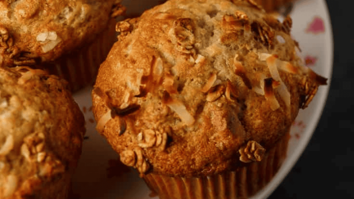 A close-up of golden-brown muffins from classic muffin recipes, topped with oats and toasted coconut flakes, arranged on a white plate with a decorative pattern.