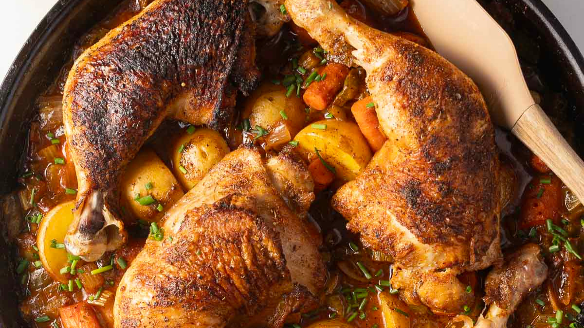 Three browned chicken legs are cooked in a skillet with golden potatoes, carrots, onions, and a rich sauce—a classic among chicken thighs recipes. A wooden spatula rests nearby, ready to serve this hearty dish.
