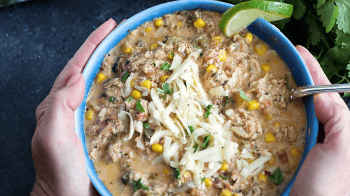 A person holds a blue bowl of creamy chicken chili, a favorite among slow cooker recipes, topped with shredded cheese, corn, and herbs and garnished with a lime wedge. A spoon rests in the bowl, with fresh cilantro in the background.