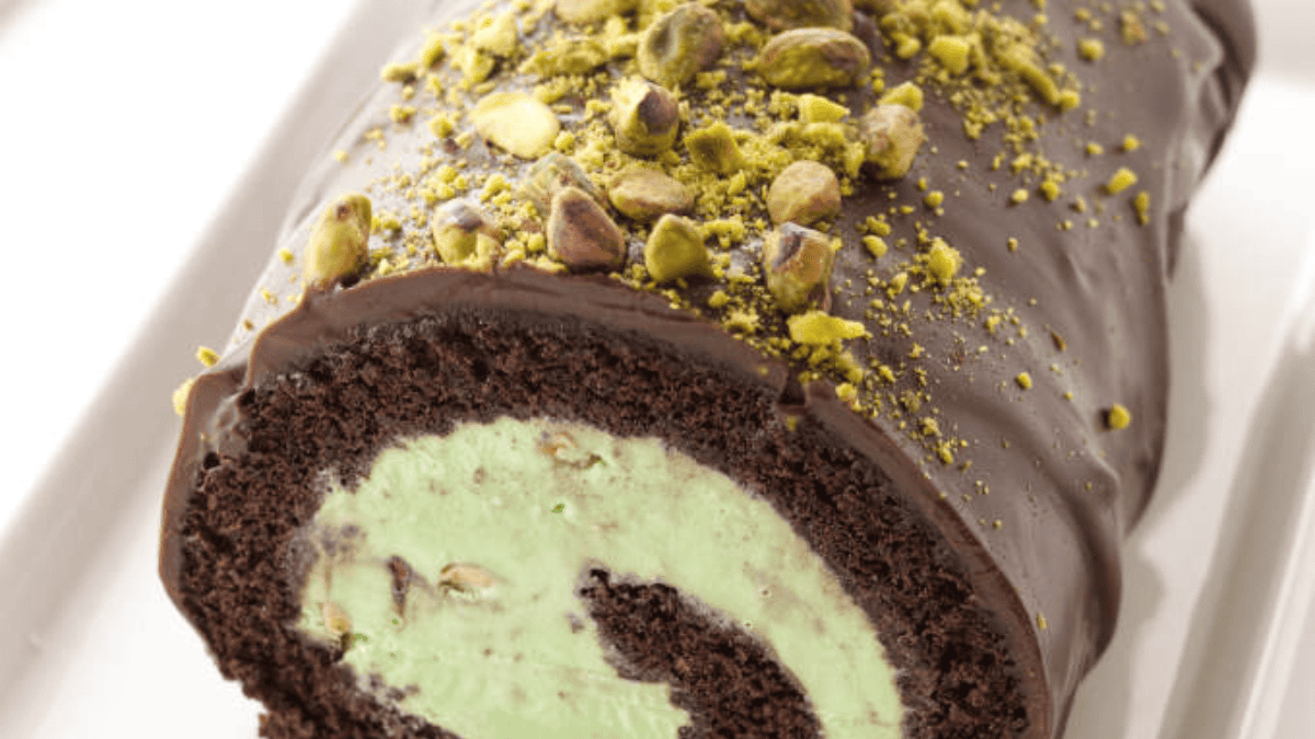 A chocolate Swiss roll cake filled with green pistachio cream, covered in chocolate glaze, and topped with chopped pistachios—perfect for fans of pistachio dessert recipes—displayed on a white plate.