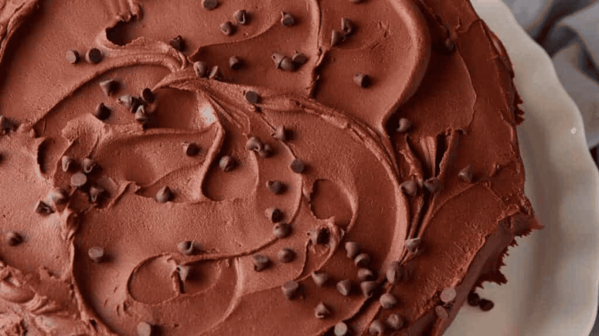 A close-up of a chocolate cake topped with swirled chocolate frosting from tried-and-true frosting recipes and scattered chocolate chips, displayed on a white plate.
