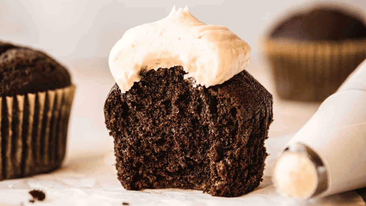 A chocolate cupcake with a bite taken out, topped with vanilla frosting. A piping bag with frosting lies nearby, and another whole cupcake—perfect inspiration for chocolate cupcake recipes—is visible in the background.