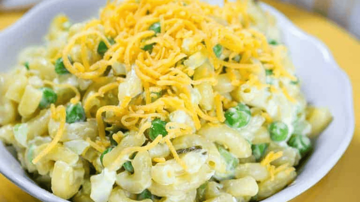 A bowl of creamy macaroni salad with green peas, diced vegetables, and topped with shredded cheddar cheese—perfect for fans of delicious pasta salad recipes.