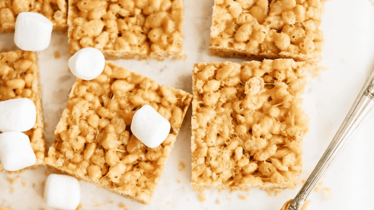 Four rice krispie treats bars and five mini marshmallows are arranged on a white surface next to a silver utensil. The treats are golden and appear chewy with visible puffed rice pieces.