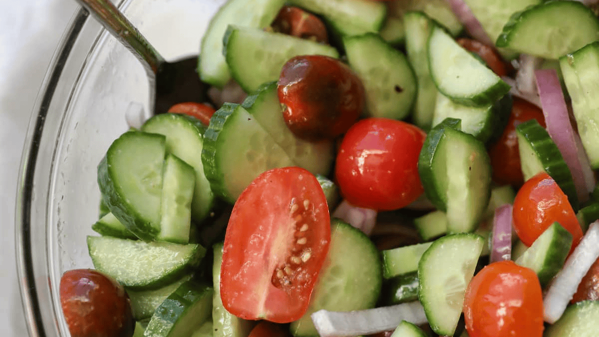 A close-up of a fresh, easy salad with sliced cucumbers, halved cherry tomatoes, red onion slices, and a spoon in a glass bowl.