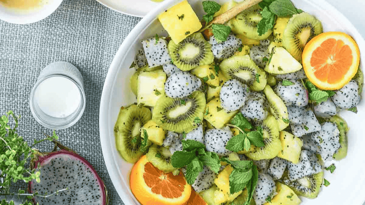 A bowl filled with a fruit salad containing sliced kiwi, pineapple, dragon fruit, fresh mint, and orange slices sits on a gray textured surface next to a small jar and half dragon fruit—perfect for lovers of colorful fruit dishes.