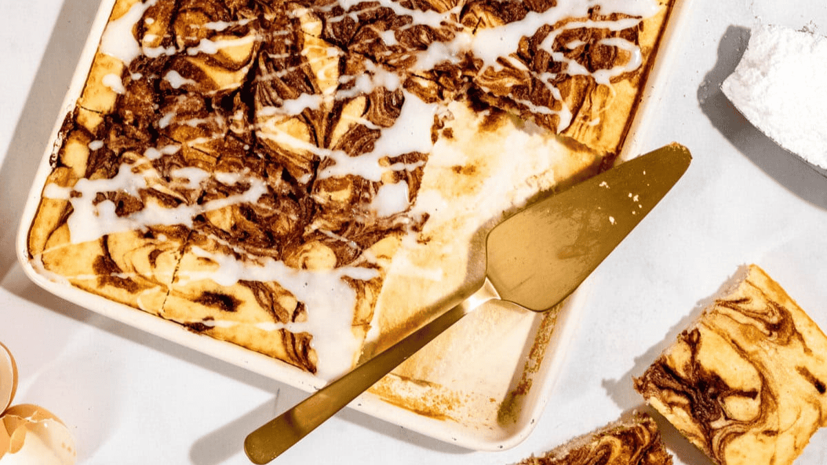 A rectangular sheet cake of marble cake with white icing drizzle, partially sliced, sits on a white surface next to a gold cake server, eggshells, and a pile of powdered sugar.