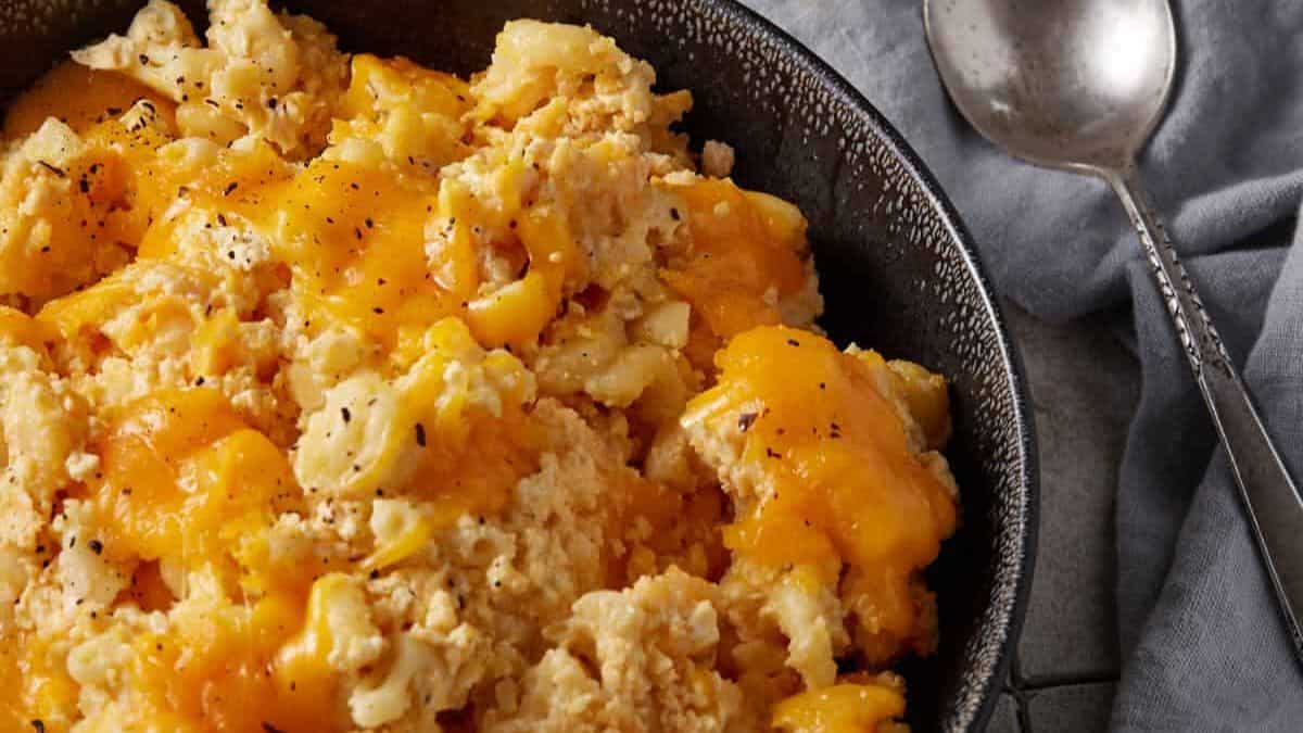 A close-up of creamy scrambled eggs topped with melted cheddar cheese, served in a black skillet next to a spoon on a gray cloth—comfort food perfect for pairing with cozy slow cooker dinners.