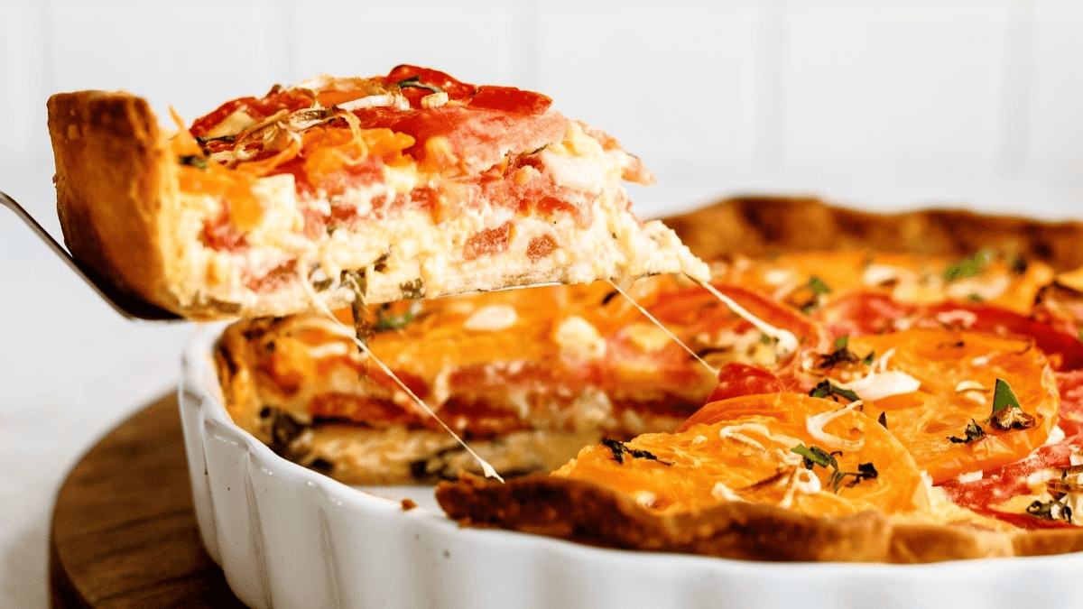 A close-up of a tomato pie in a white dish, with a slice being lifted out, showing layers of tomatoes, cheese, and herbs in a golden, flaky crust—a true nod to southern comfort food classics.