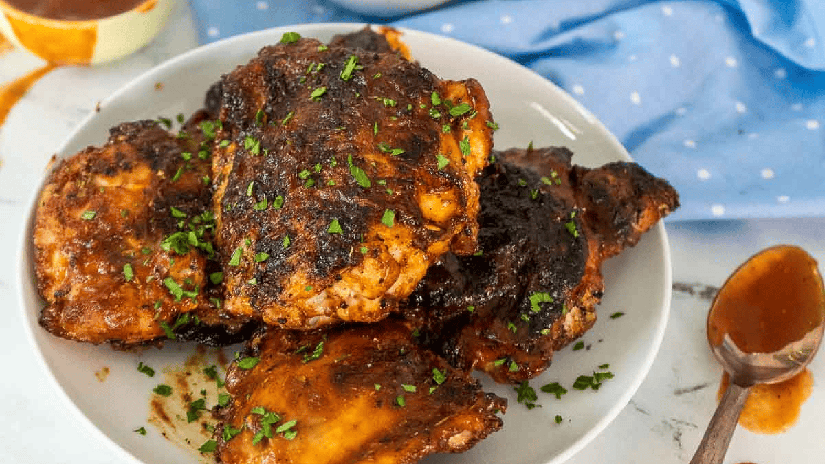 A plate of grilled chicken thighs, inspired by classic chicken thighs recipes and coated in a dark barbecue sauce, is garnished with chopped herbs and sits on a white table next to a spoon and a blue polka-dot cloth.