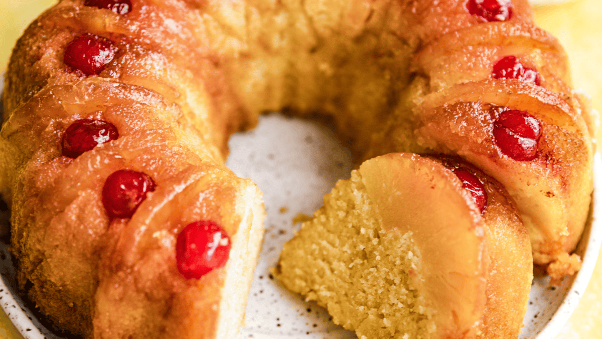 A close-up of an easy bundt cake topped with pineapple rings and red cherries, with a slice cut out and resting on a speckled plate. The cake looks moist and golden with a caramelized top.