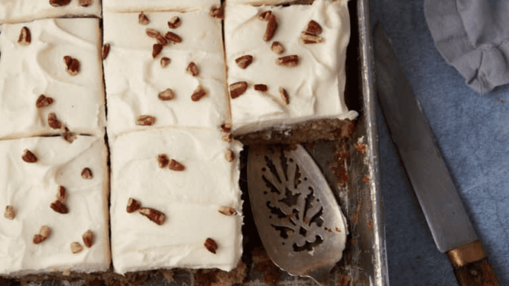 Tray of sliced sheet cake topped with creamy white frosting and sprinkled with chopped pecans, with a serving spatula partially under one piece and a knife resting beside the tray.