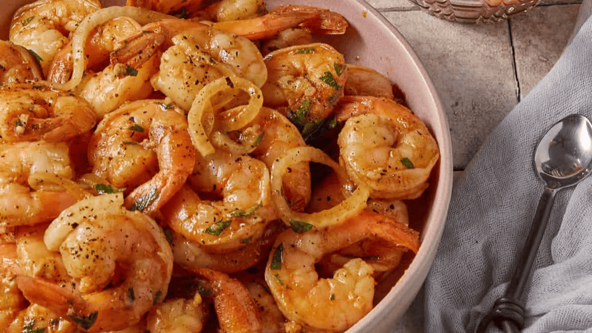 A bowl filled with cooked shrimp garnished with herbs and thin onion rings, seasoned with pepper and inspired by classic shrimp recipes, sits on a tiled surface beside a gray napkin and a spoon.