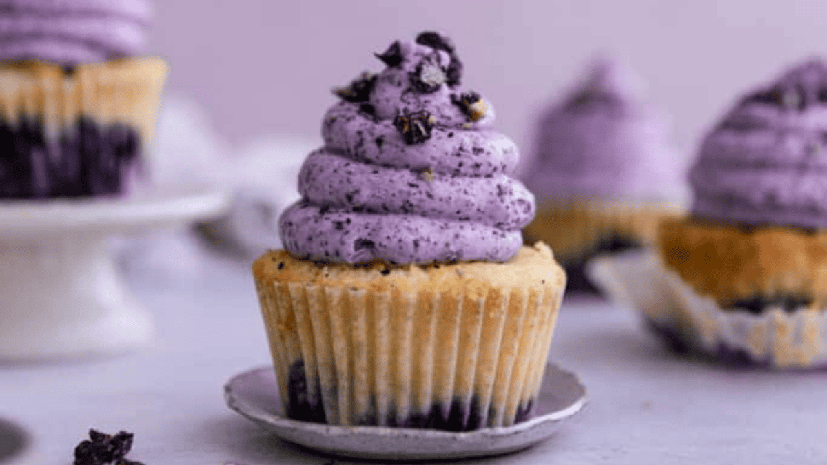 A blueberry cupcake topped with swirled purple frosting and blueberry pieces, placed on a small plate with more cupcakes in the background—perfect inspiration for your next batch of blueberry recipes.