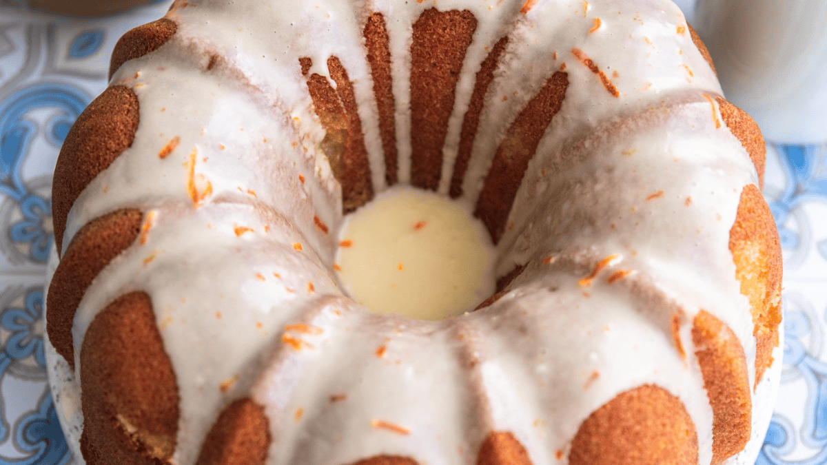 An easy bundt cake covered in white glaze sits on a blue and white patterned surface. The glaze drips down the sides and is topped with small pieces of orange zest.