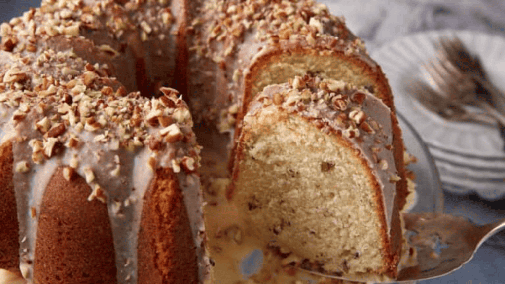 A sliced easy Bundt cake topped with glaze and chopped nuts sits on a plate, with a cake server lifting one slice. Stacked plates and forks are in the background.