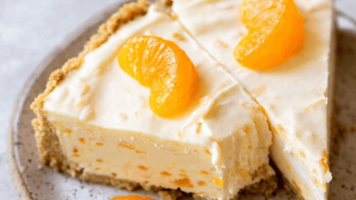 A creamy no bake dessert pie with a crumbly crust, filled with a light orange mixture and topped with mandarin orange slices, served on a speckled plate.