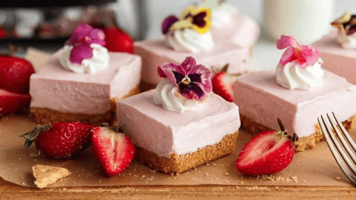 Squares of pink no bake cheesecake with graham cracker crust are topped with whipped cream and edible flowers, arranged on a wooden board with halved fresh strawberries scattered around. Perfect for fans of no bake desserts.