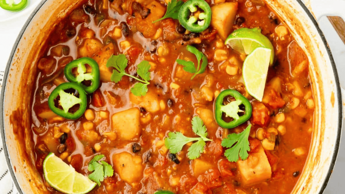A pot of chili filled with beans, corn, tomatoes, potatoes, and spices—an affordable dinner—garnished with lime wedges, fresh cilantro, and slices of jalapeño pepper.