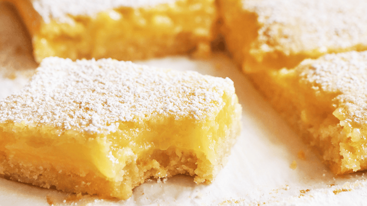 Close-up of lemon bars with a golden shortbread crust, topped with powdered sugar. One bar in the foreground has a bite taken out, revealing creamy lemon filling—an irresistible treat for lovers of classic shortbread recipes.