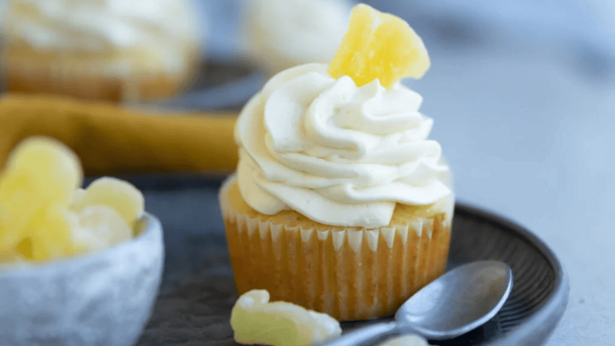 A cupcake with swirled white frosting and a small pineapple slice on top sits on a dark plate next to a spoon, with a bowl of pineapple chunks in the background—perfect for fans of quick desserts.